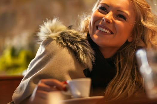 Amidst a quaint European café, a young lady indulges in a sun-kissed espresso. Winter's grasp evaded, clad in fur, exquisite solitude, yet open to chance encounters