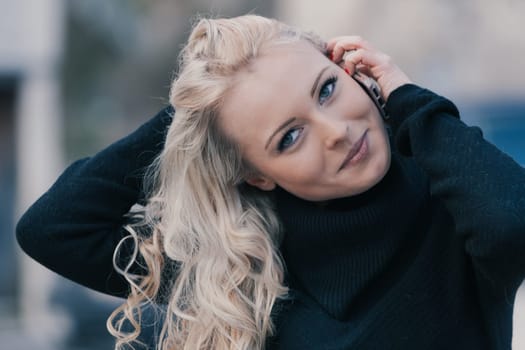 Young woman with long, curly, perfectly permed blonde hair, perfect makeup and manicure, is a tourist in a European city with medieval villages. Using her phone to talk, chat, or access Wi-Fi for hist