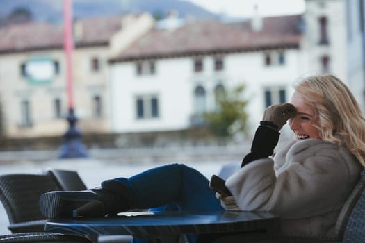 Beautiful young blonde woman sitting at a bar, having fun, laughing at something hilarious on her smartphone or a received message. She's a tourist in a European city, enjoying the fresh air, dressed