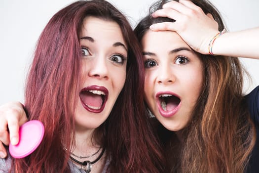 Two youthful friends, impeccably made-up and with flowing hair, let loose, making funny faces. They revel in the carefree moments and their playful expressions shine through