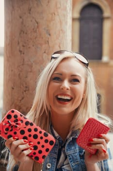 With a European city's historic architecture as her backdrop, a blonde girl enthusiastically shows two pink wallets. Her facial expression screams shopping delight as she plans to dip into her saved f