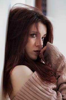 Sensual young woman leans against a wall. Red hair cascades, strand touching lips. Her pink sweater reveals a shoulder her gaze and dark lipstick convey confidence and allure