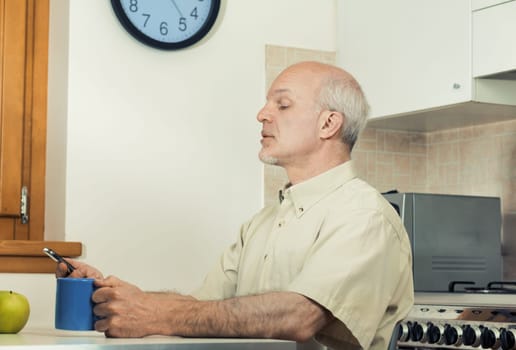 Elderly man struggles with his smartphone in the kitchen. Modern phones don't cater to those with poor vision. Attempting breakfast, he squints, seeing blurry. An eye check and glasses might help