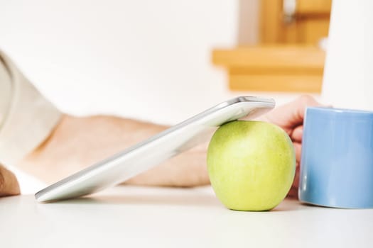 In a symbolic setup, a digital tablet stands on a green apple, accompanied by a blue mug in a kitchen. The presence of a man's arm, face not visible, indicates a motive: to utilize these tools to bene