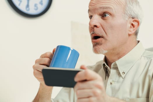Elderly man in his kitchen looks at a smartphone with great astonishment. The news or videos he views oppose tradition or alarm him. His morning coffee might choke him from indignation