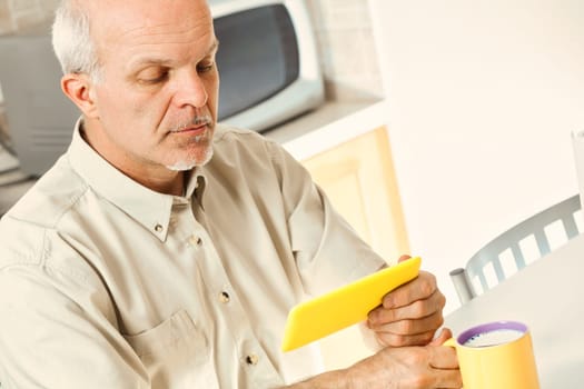In his kitchen, a senior finds using his smartphone challenging. He squints over breakfast, indicative of phones not designed for vision issues. Perhaps glasses could assist him