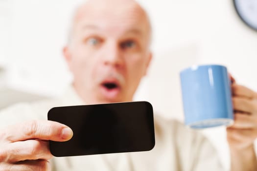 In the kitchen, an old man experiences shock from viewing content on his smartphone. The materials, clashing with tradition or causing alarm, could upset his morning coffee ritual