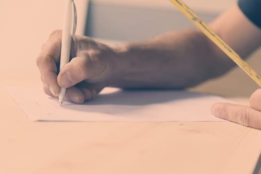 hand of a wood craftsman writing with a ballpoint pen on a paper resting on a rough-hewn wooden board while holding a tape measure with the other. He takes measurements and makes an outline drawing