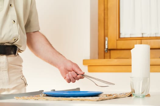 In the intimate setting of his kitchen, an unidentified man's arm reaches out. He's setting the table, laying a knife and fork adjacent to the plate. His neat attire implies a solitary existence