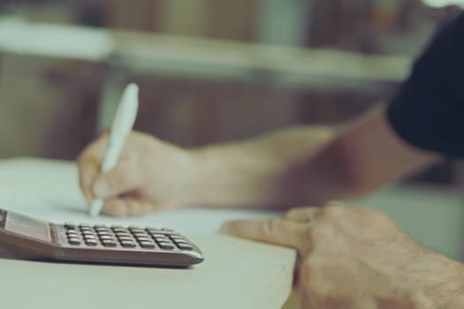 A man's hands blurred as they write on a paper resting on a table. In the foreground, in focus, a pocket calculator. Craftsman at work