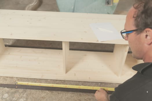 Carpenter takes measurements of a piece of furniture with a tape measure in his craftsman's workshop