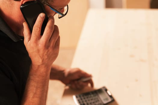 wide copyspace on the lighted table of a carpenter making estimates with a calculator while hearing the supplier on the phone. He's wearing glasses.
