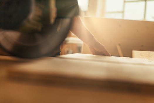 The carpenter guides a wooden board with a strong and confident hand as it is cut by a circular saw. Wood chips and sawdust fly through the air as the blade whirls, illuminated by the sunlight in his