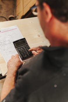 Craftsman or worker performs calculations with a pocket calculator leaning against a wooden shelf in the background in focus, while he is out of focus in the foreground.
