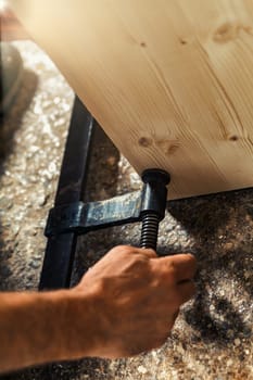 The carpenter's hand secures two parts of a furniture piece with a wooden screw clamp in his artisanal workshop, illuminated by warm sunlight pouring through the window