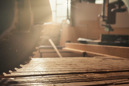 The circular saw is stationary in the carpentry shop, the sun's rays piercing through the sawmill workshop window striking the razor-sharp teeth and grooves of the wooden boards. Romantic and dangerou