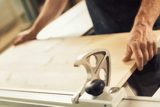 n a woodworking shop, a man is positioning a wooden shelf or board onto a lumber processing machine. Strong male hands and large amounts of wood are visible, as well as the metal structures formed by