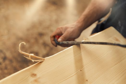 The artisan carves and chamfers the edges of the wooden boards that make up the furniture with a drawknife. Strong male hands and wood are visible, embodying the essence of carpentry