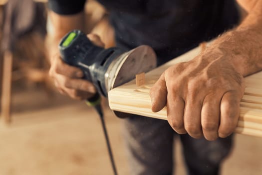 The carpenter's hands grip a sander. The wood artisan, an experienced professional, holds a board or piece of wood with strong and precise hands, carefully sanding it with a machine that vibrates sand