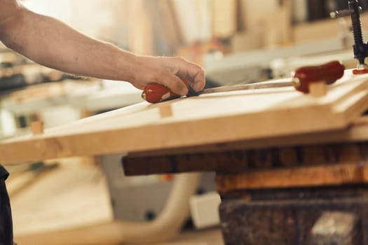 n a sawmill or artisanal carpentry workshop, a carpenter's hand grips the handle of a drawknife resting on light-colored wood. The artisan has strong and virile arms