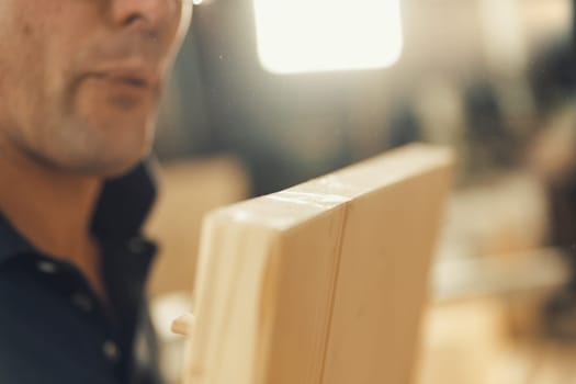 In a shallow depth of field shot, the carpenter blows sawdust off a freshly cut piece of wood, holding it firmly with both hands, while the rest of the workshop is blurred