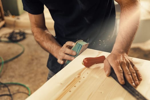 The artisan is completely hand sanding with one hand, while the other rests on a wooden surface holding a drawknife used for artistic beveling