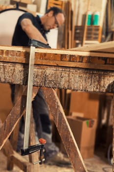 Foreground features a wooden clamp hanging from a workbench on sawhorses, while in the blurred background, the bespectacled carpenter works in a traditional workshop that modernizes daily