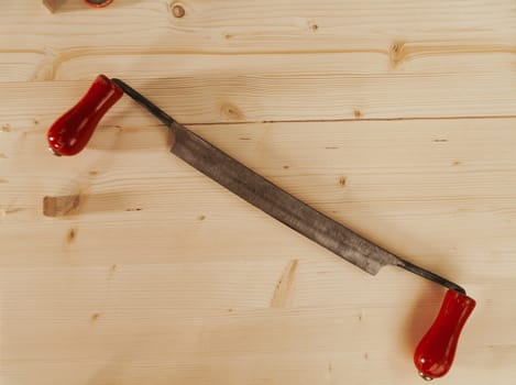 Top view shot of a carpenter's hand cutting tool. The drawknife has smooth red handles and is resting on light-colored wood.
