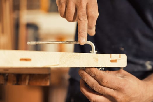 The carpenter screws and secures a wooden board using an Allen key, holding the other side with one hand. Strong, muscular, and hairy arms and hands are visible, exemplifying the manual labor involved