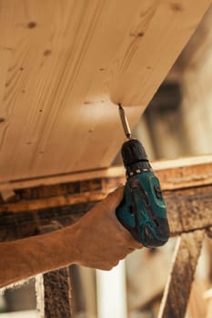 Carpenter's hand using an electric screwdriver with illuminated tip under a shelf on wooden sawhorses. His strong, steady hand is precise and controlled. Detail in focus, the rest is blurred
