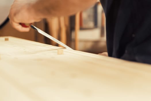 An artisan stands with his drawknife in hand. This carpenter loves to use his manual cutting tool because he desires control and the ability to rely solely on his own hands. In the background, his wor
