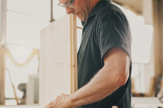 Carpenter strives in a movement that needs precision but also muscle power. He wears glasses and stands in his beautiful carpentry shop lit by the sun coming in through the large window.