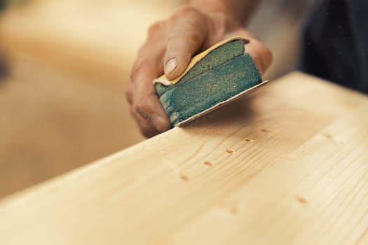 A close-up shot of the carpenter's hands using sandpaper to manually sand an axis, while resting it on his work surface. The rest of the shot is blurred