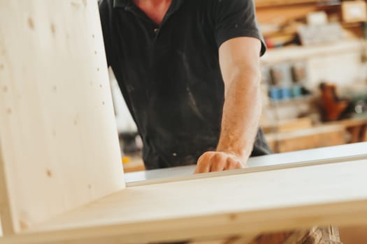 Carpenter's hand pushing and supporting a piece of furniture under construction. Blurred in the background is his artisan carpenter's workshop. Strength and determination are his main tools.