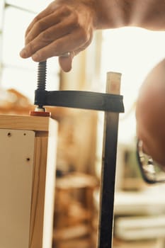 Wood artisan works traditionally in his shop. Carpenter tightens a wooden screw clamp to secure two handmade furniture parts in his brightly lit workshop. Wood, light, strength