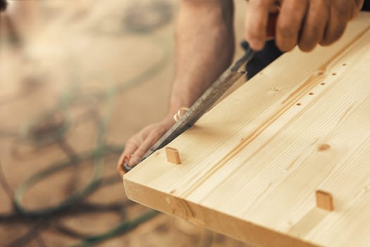 The artisan carves and chamfers the edges of the wooden boards that make up the furniture with a drawknife. Strong male hands and wood are visible, embodying the essence of carpentry