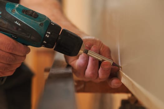 In focus: cordless drill-driver with built-in LED light in the hands of an experienced carpenter. He is located in his own workshop and uses every necessary tool.