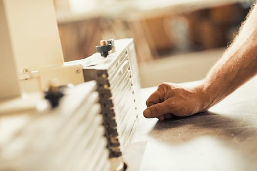 A carpenter's strong and muscular hand and forearm holding a metal tool while working on a self-operating wooden piece of furniture. The warm sunlight flooding the workshop and the wood