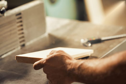A carpenter's strong and muscular hand and forearm holding a metal tool while working on a self-operating wooden piece of furniture. The warm sunlight flooding the workshop and the wood