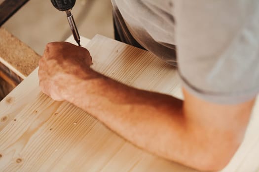 Detail in focus on the action of screwing a hinge onto a handcrafted piece of furniture produced by a carpenter. Strong hands and arms of the craftsman in view and lots of light wood.
