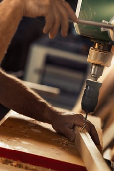 Carpenter uses drill press in his workshop. With his left hand, taking calculated risks, he holds and guides the wood, while lowering the lever with his right hand to control the exact spot and pressu