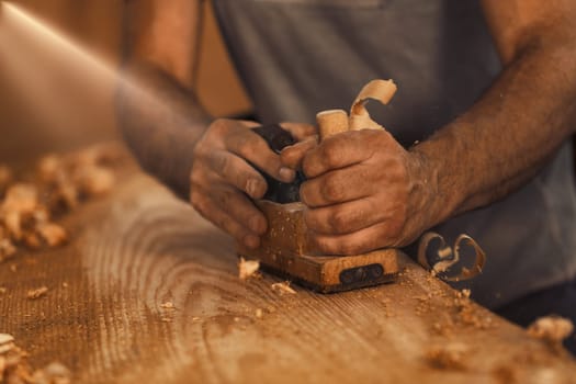 The wood artisan, a robust and strong man known as a carpenter, hand planes a board in the traditional manner, producing shavings of sawdust that look like golden curls of wood. It is a strenuous and