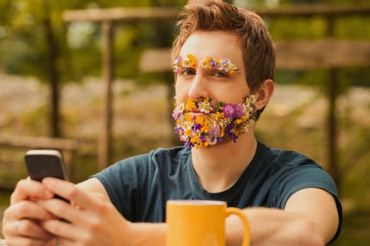 Young man with colorful spring flowers instead of facial hair sits amidst nature. While cherishing the tranquility and fresh air, he doesn't despise technology, staying connected via phone