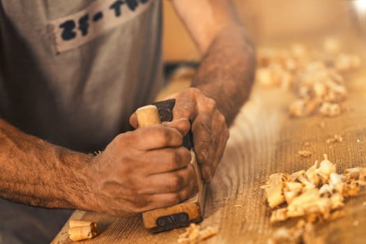 The wood artisan, a robust and strong man known as a carpenter, hand planes a board in the traditional manner, producing shavings of sawdust that look like golden curls of wood. It is a strenuous and