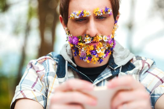 With a beard and eyebrows made of vibrant spring blooms, a man sits in nature's lap. He stays in touch through his phone, embracing both peace and technology