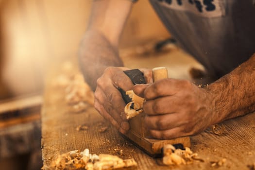 The wood artisan, a robust and strong man known as a carpenter, hand planes a board in the traditional manner, producing shavings of sawdust that look like golden curls of wood. It is a strenuous and