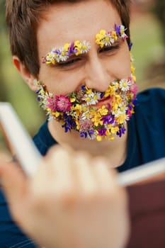 Amidst trees, a unique sight unfolds: a young man's facial hair blooms as vibrant spring flowers. He's engrossed in a traditional leather-bound tome