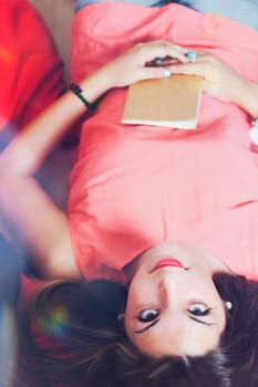 Reversed shot of young woman lying with a notebook on her belly and idyllic 1970s lighting overlays, she has long brown hair and looks into the camera