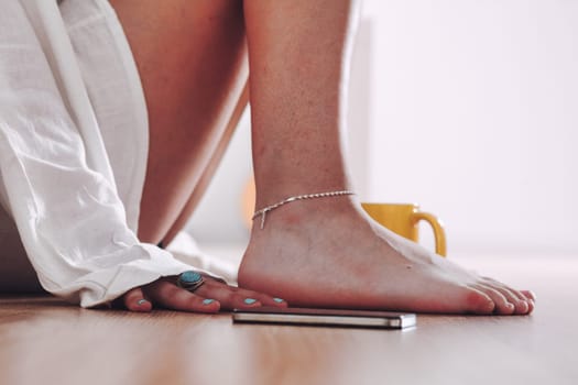 Close-up shot, detail, of huddled woman's or girl's legs, of her bare feet with bracelet. She wears a loose white shirt and is sitting on the floor, her hand with rings on the ground still near her sm