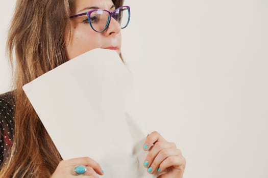 woman with very long old-fashioned brown hair and very wide glasses wears a dark dress with light semi-transparent dots. She nibbles nervously at the corner of the paper she holds in her hands while l